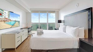 A hotel room with a large bed, a desk, a TV, and a balcony with a view of a building and a pool. - Coconut Grove Suite | 20th Floor Bay Views (Miami)