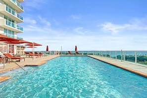 Ein Swimmingpool mit Liegestühlen und Sonnenschirmen, umgeben von einem Hochhaus mit Balkonen.
