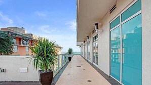 Ein langer, schmaler Balkon mit Blumentöpfen, blauem Himmel und Gebäuden im Hintergrund.