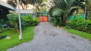 Un chemin de gravier mène à un portail en bois, entouré d’une végétation luxuriante et d’une clôture en métal.