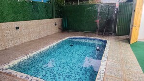 Une piscine rectangulaire bordée de mosaïques, entourée d’un mur de pierre et d’une clôture verte.