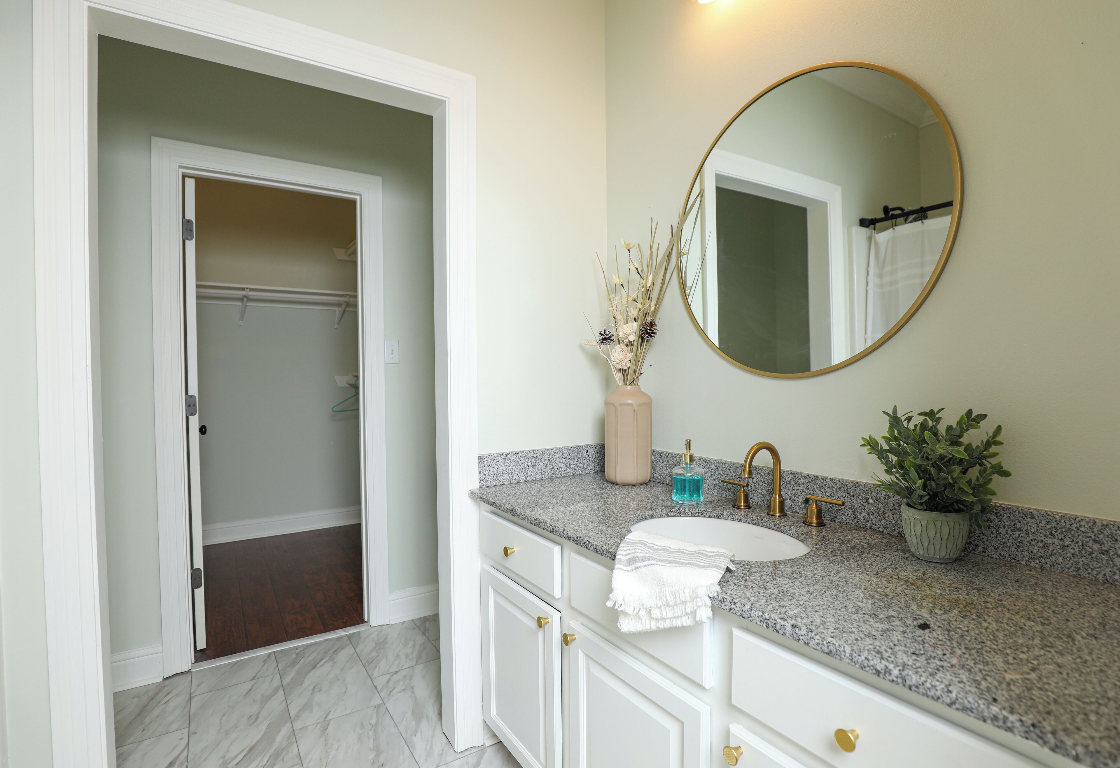 Une salle de bain avec un comptoir en granit, un lavabo blanc, un miroir rond, une serviette et une plante en pot.