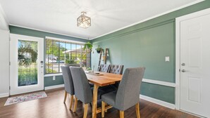 Een eetruimte met een houten tafel, grijs gestoffeerde stoelen, een hanglamp en uitzicht door een glazen deur.