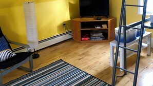 Huoneessa on keltaiset seinät, kerrossänky, puinen tv-taso, jossa on televisio, raidallinen matto ja tyynyillä varustettu tuoli.