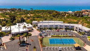 Luchtbeeld van een hotel met een tennisbaan, parkeerplaats en uitzicht op de oceaan.