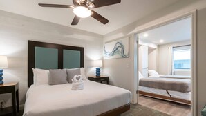 Hotelkamer met een groot bed, een plafondventilator, nachtkastje met lamp en uitzicht op de aangrenzende kamer.