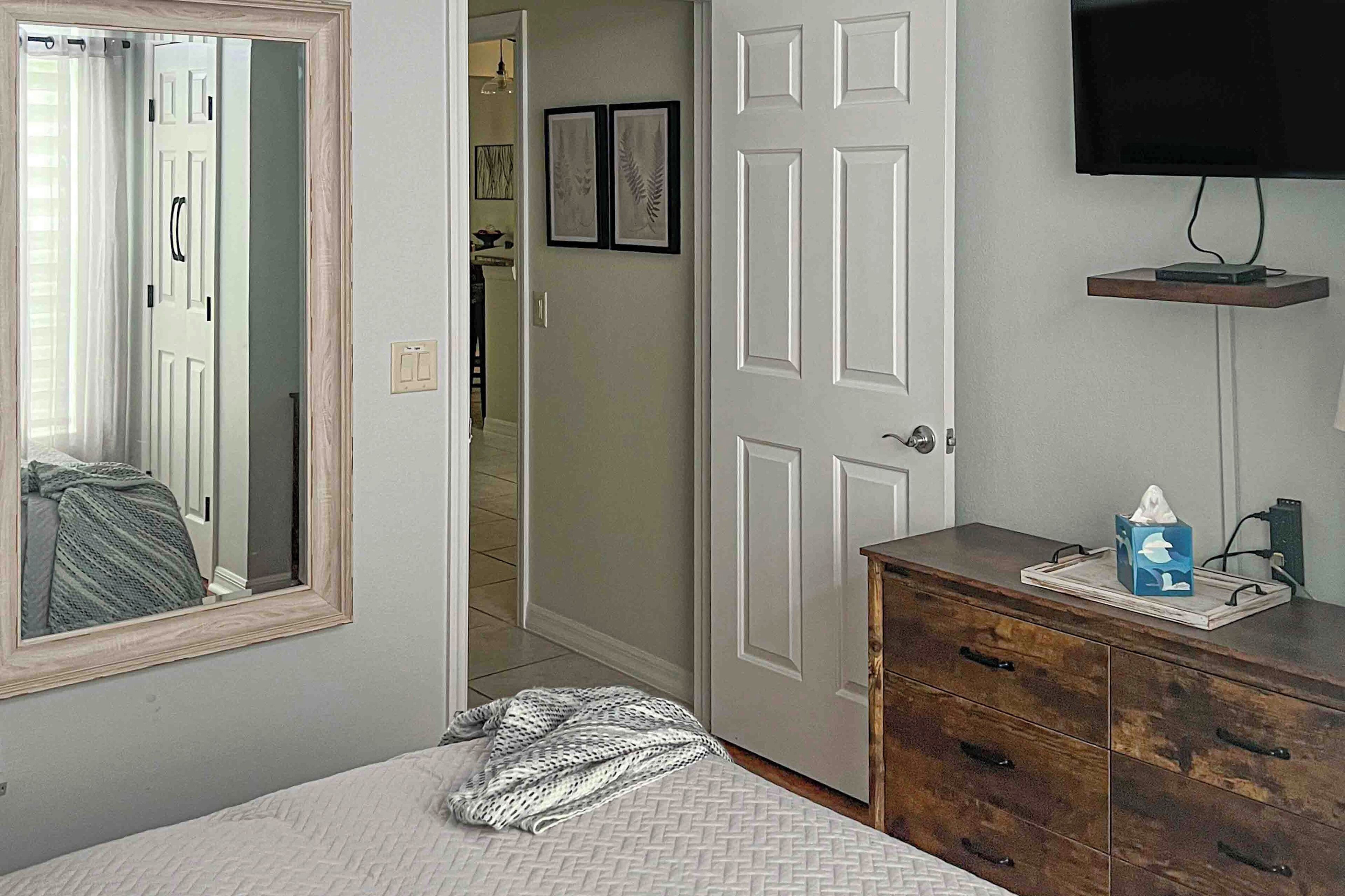 Un dormitorio con cama, cómoda, espejo, puerta y televisor montado en la pared.