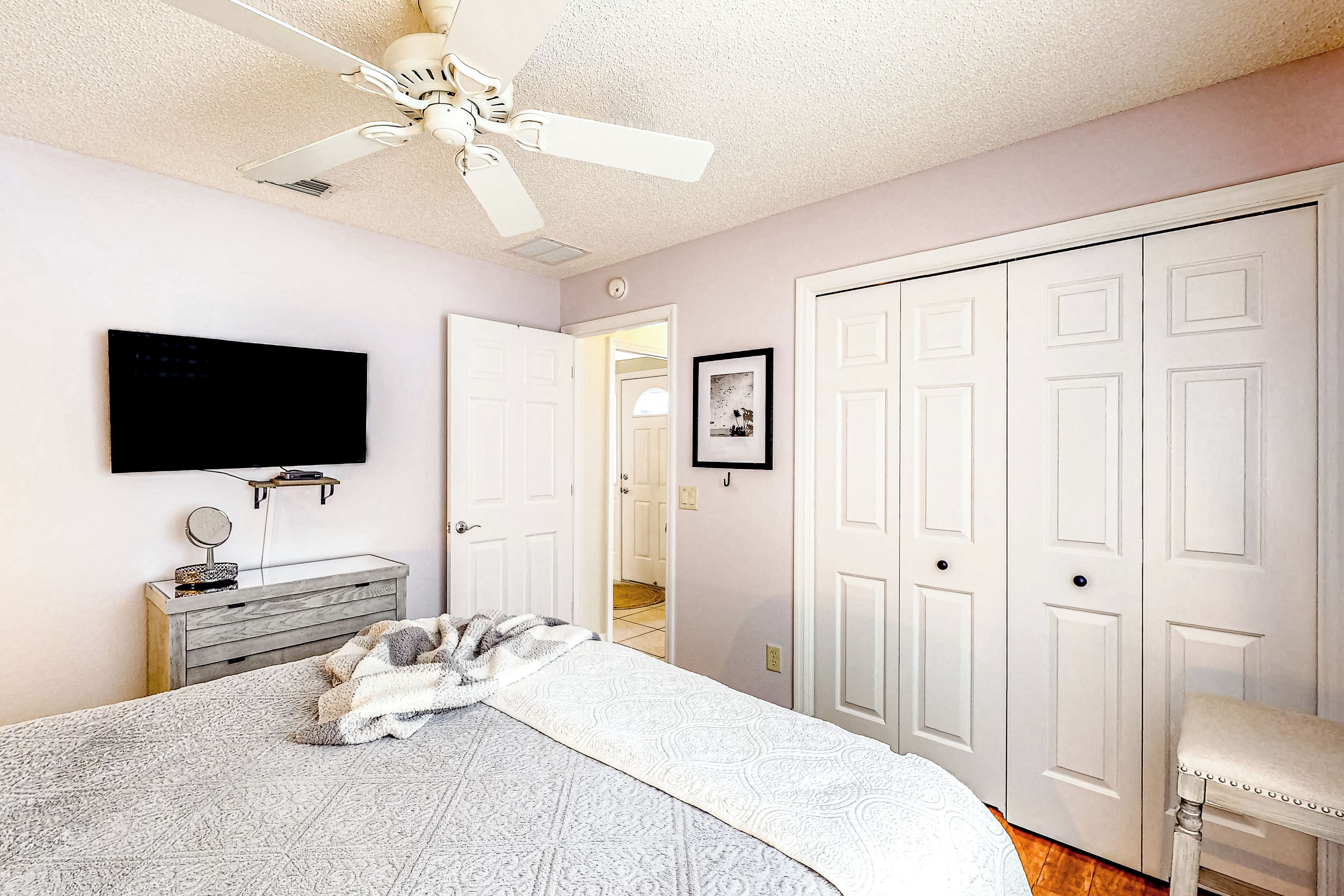 A bedroom with a bed, a ceiling fan, a TV mounted on the wall, a dresser, a closet, and a door leading to another room.