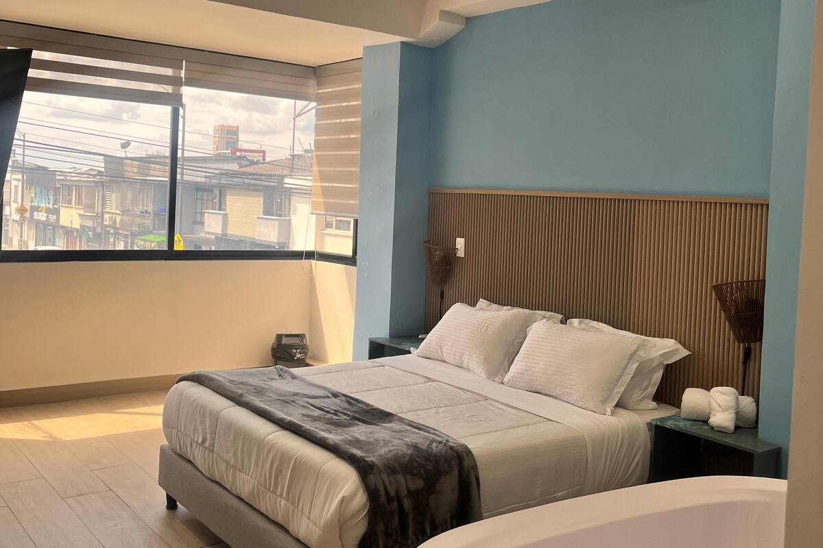 A modern bedroom with a large bed, a nightstand, a bedside table, a window with blinds, and a view of buildings outside.