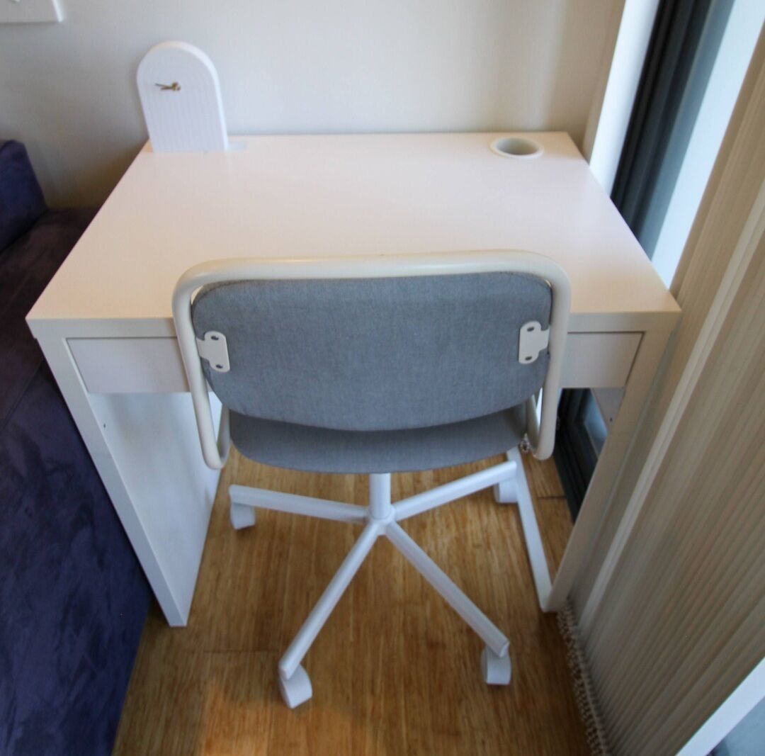 A compact, white desk with a grey cushioned chair in a room with wooden flooring and a window.