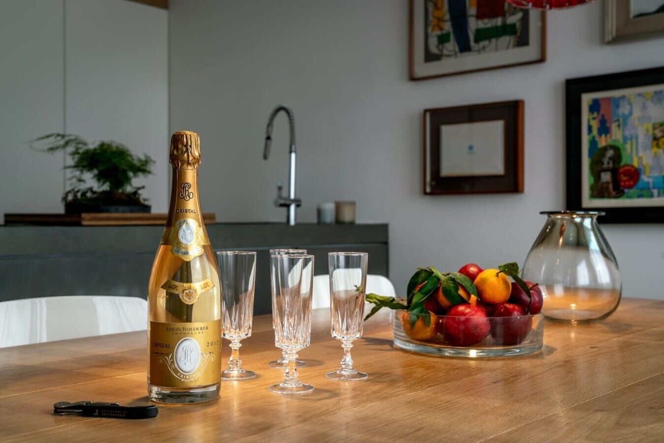 A bottle of Cristal champagne, four glasses, a fruit bowl, a key, and a vase on a wooden table.