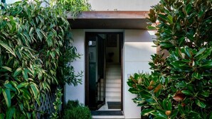 Une entrée moderne, avec une porte en verre, un chemin en pierre et une végétation luxuriante de part et d’autre.