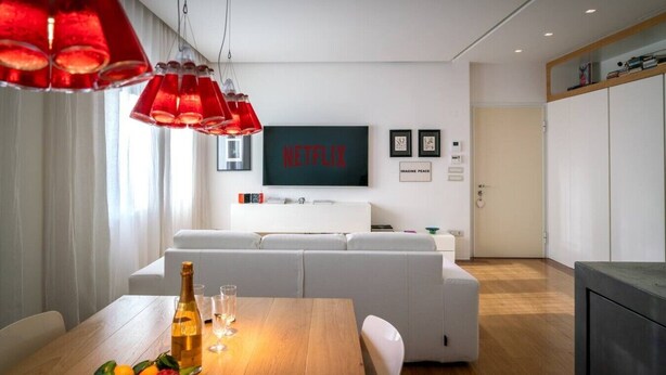 Un salon moderne avec un canapé blanc, une table à manger en bois sur laquelle sont disposés des fruits et des verres, une suspension rouge et un téléviseur mural qui affiche le mot « NETFLIX ».