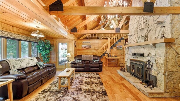 Acogedor interior de cabaña de madera con chimenea, sofá de cuero y una escalera que lleva a un nivel superior.
