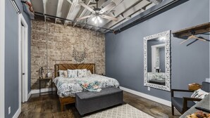 A bedroom with a brick wall, a bed with floral bedding, a grey bench, a mirror with an ornate frame, and a ceiling fan. - Downtown Urban Loft above Restaurants (Perryville)