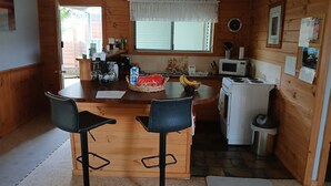 A compact kitchen with a central island, two bar stools, a microwave, a refrigerator, and a window with a white curtain. - Snug Stays After Wild Days (Strahan)