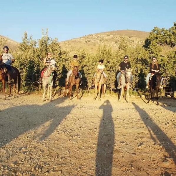 A group of people riding horses on a dirt path with sparse vegetation and a hilly background.