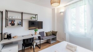 A modern, well-lit room with a white desk, a comfortable seating area with cushions, a mounted TV, and a large window with sheer curtains. - Batèl House 5 (Como)