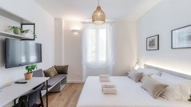 A modern bedroom with a bed, bedside tables, a desk, a TV, and a hanging light fixture. - Batèl House 5 (Como)