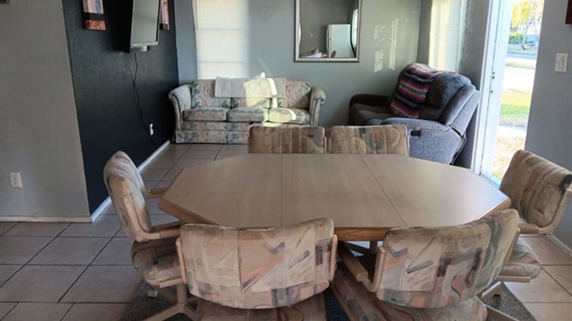 A dining area with a large wooden table and patterned chairs, a sofa, and a window with blinds.
