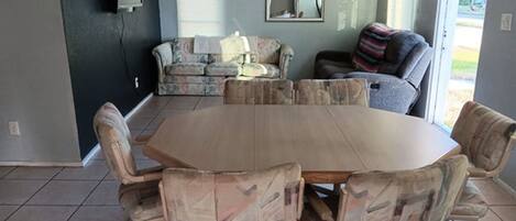 A dining area with a large wooden table and patterned chairs, a sofa, and a window with blinds.