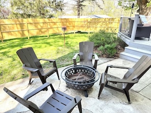 A patio area with four Adirondack chairs, a fire pit, and a wooden fence in the background.