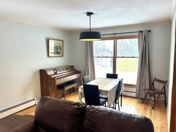 Una sala de estar con un piano, una mesa de comedor con sillas, un sofá y un ventanal con cortinas.