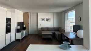 A modern, compact living space with a kitchenette, a sofa, a dining area, and a large window providing natural light. - Modern and bright home by Prime Tower with workspaces & balcony (Zürich)