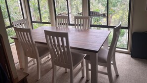A dining area with a wooden table and white chairs, large windows with a view of greenery outside.