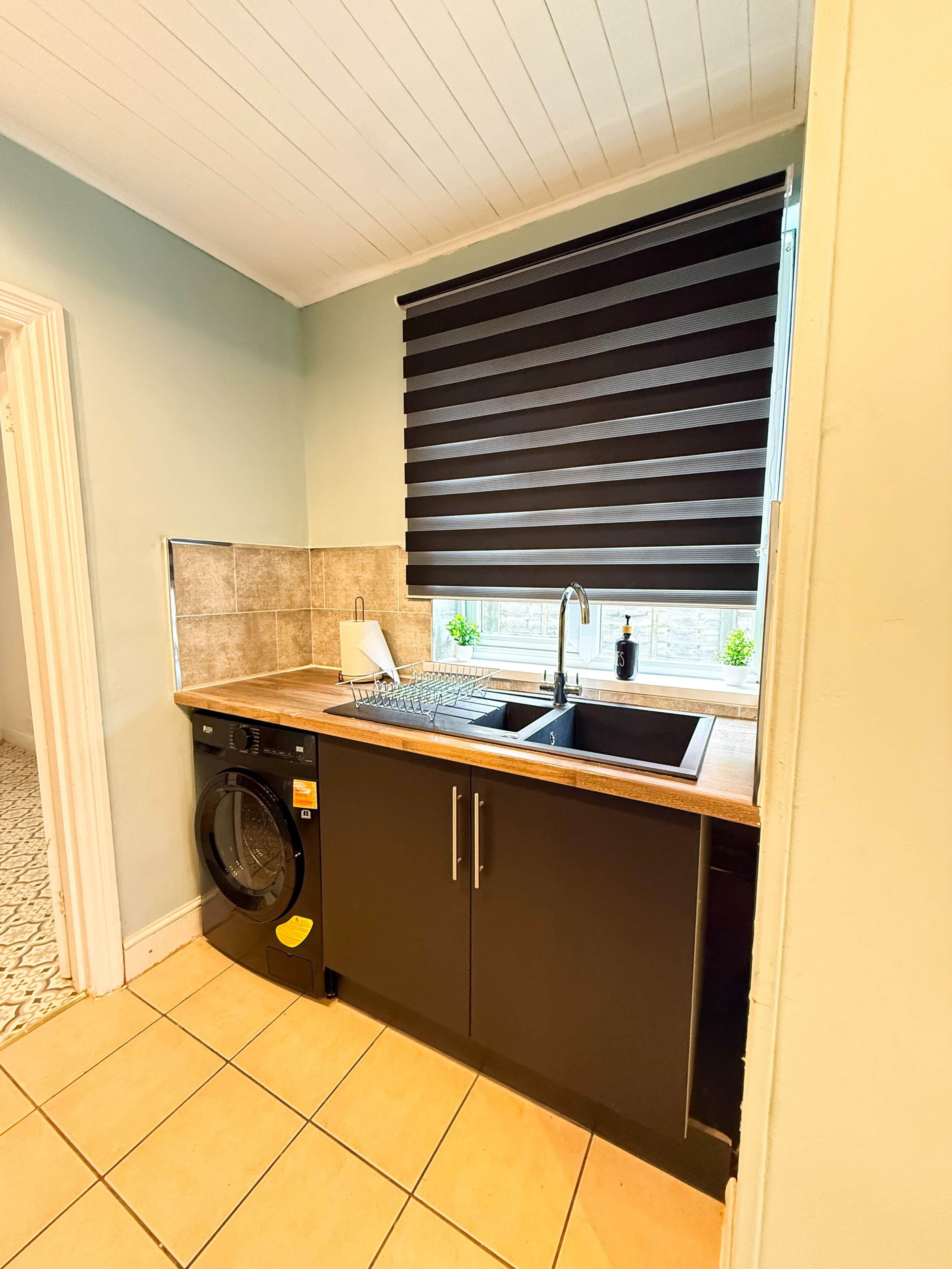 Ett kompakt kök med tvättmaskin, diskho och ett fönster med persienner.
