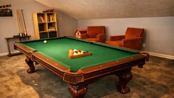A pool table with a cue ball and a rack of billiard balls, surrounded by two leather armchairs and a bookshelf.