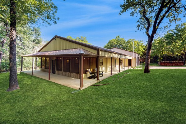 A single-story house with a covered patio area, a lawn, and trees in the background.