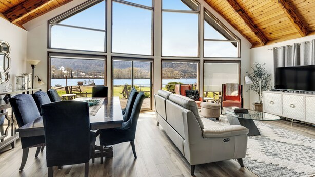 Un salón espacioso con un gran ventanal con vistas a un lago, una zona de comedor con sillas oscuras, un sofá gris y un mueble de entretenimiento moderno.