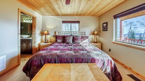 Une chambre avec un plafond en bois, un grand lit avec une literie à motifs, deux lampes de chevet, une fenêtre donnant sur un beau paysage et une table en bois placée au centre de la pièce.