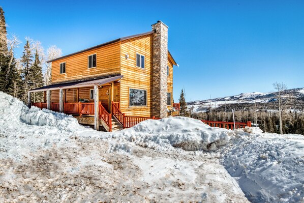 Une cabane en rondins avec une cheminée en pierre, une terrasse en bois et une vue sur des montagnes enneigées.