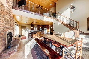 Une salle à manger spacieuse avec un haut plafond en bois, un escalier doté de rampes décoratives, et une vue sur la cuisine en arrière-plan.