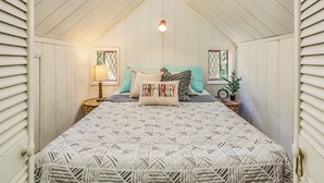 Um quarto de sótão aconchegante com teto inclinado, uma cama com roupa de cama estampada, duas mesas de cabeceira com abajures e uma janela com vista.