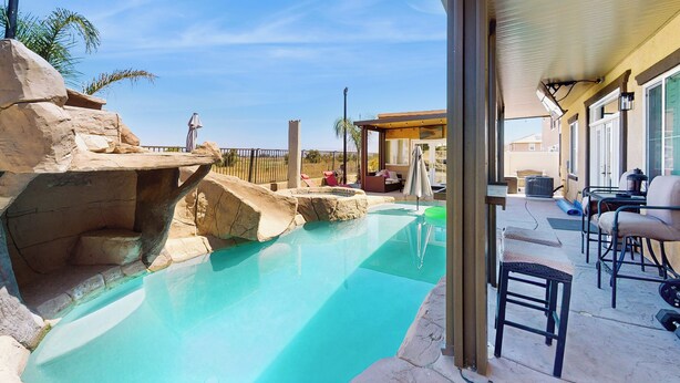 Un espace piscine agrémenté d’une formation rocheuse, de chaises longues et d’une terrasse couverte.
