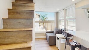 Un intérieur moderne avec un escalier en bois, un canapé gris, un bureau blanc et de grandes fenêtres donnant sur l’extérieur.