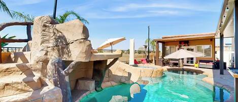 Zona de piscina con paisaje rocoso, una piscina azul cristalina y un edificio con asientos al aire libre.