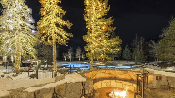 A stone fire pit surrounded by snow, with illuminated trees and a pool in the background. - Valley-View Unit in Everline Resort: Premier Perks (Olympic Valley)