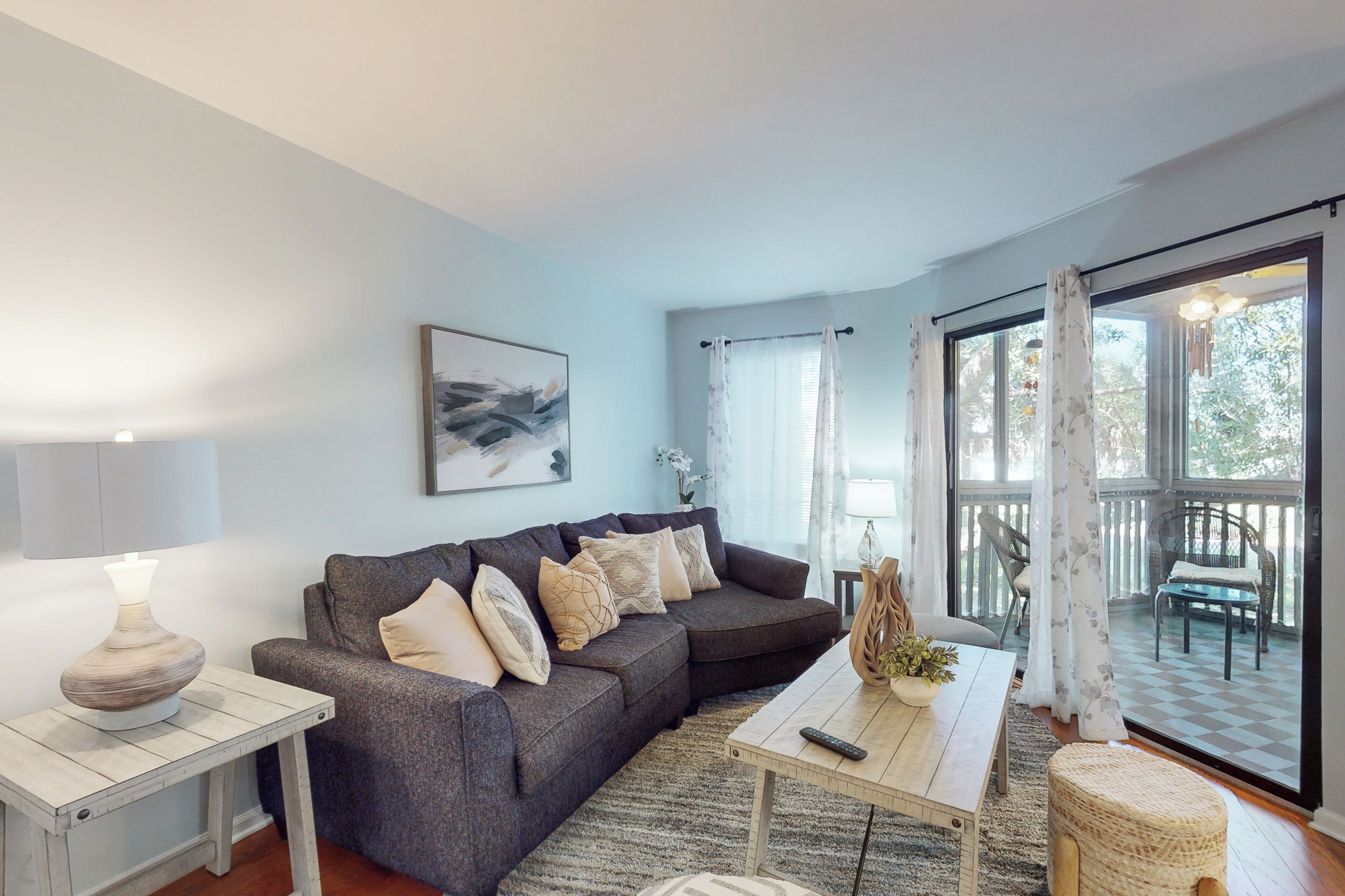 Ein modernes Wohnzimmer mit einem grauen Ecksofa, einem kleinen Holztisch, einem weißen Beistelltisch mit Lampe und Blick ins Freie durch große Fenster.