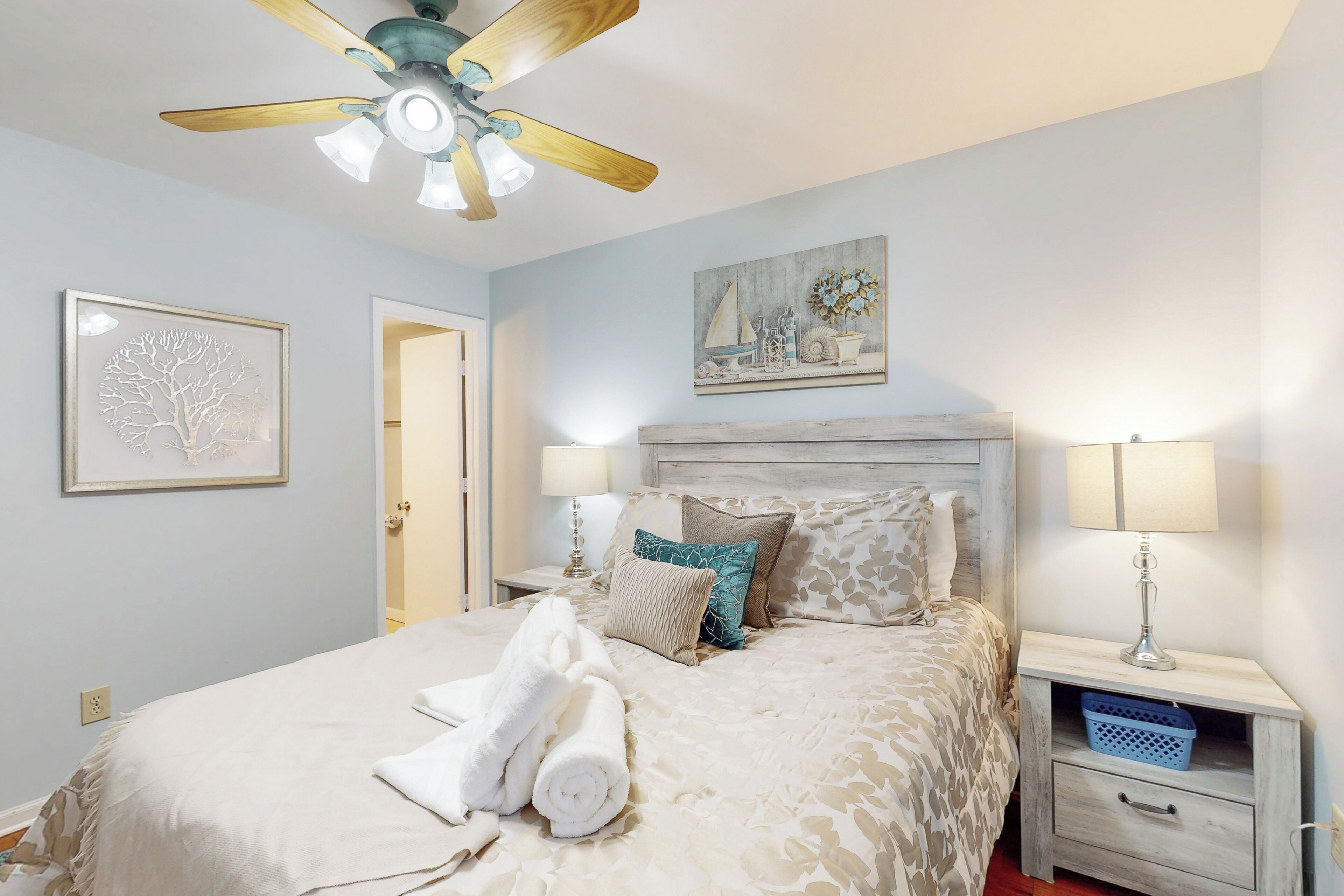 A neatly made bed with a patterned bedspread, a ceiling fan, a wall-mounted painting, and a bedside table with a lamp and a basket.