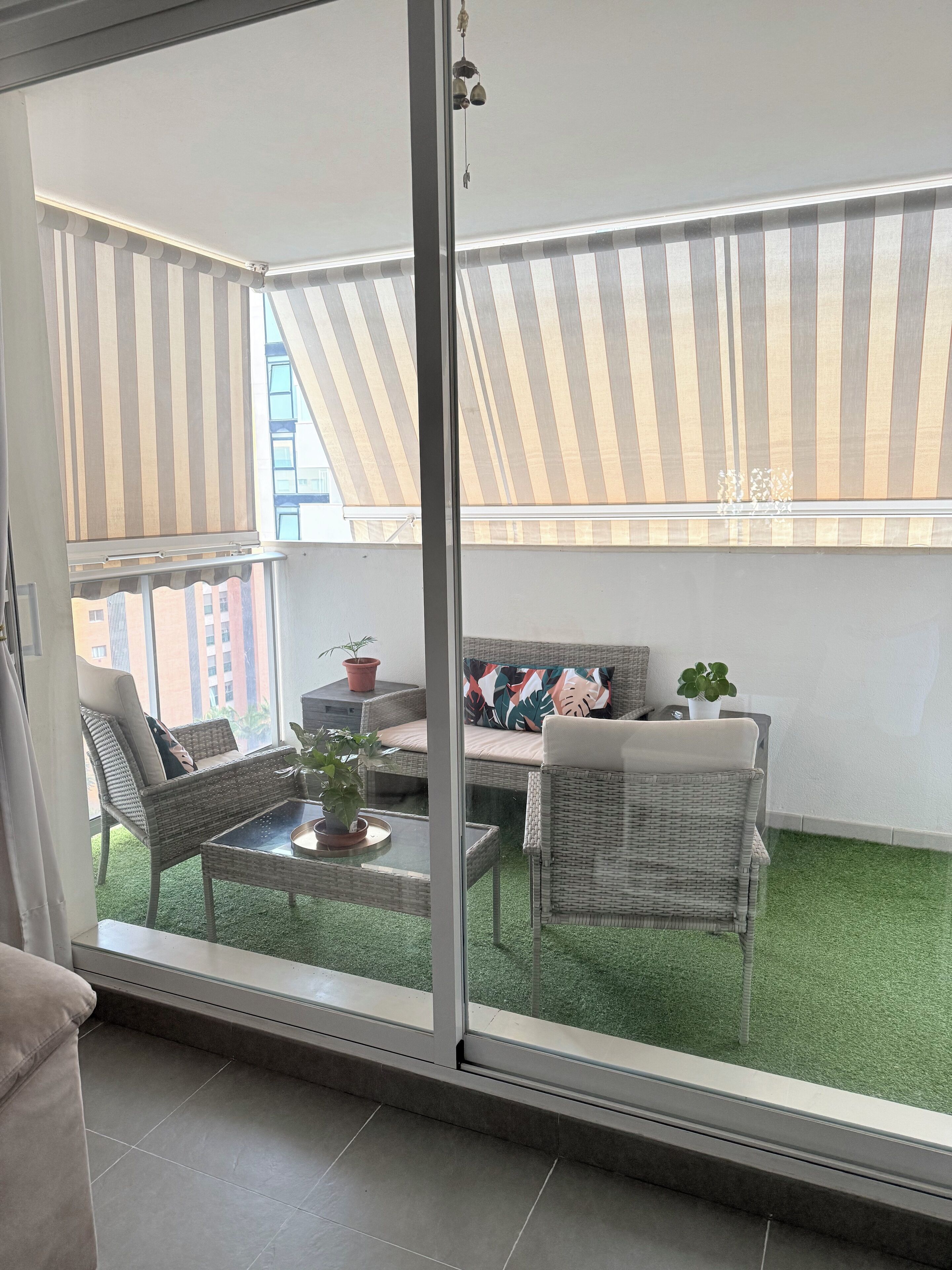 A balcony with a glass door, wicker furniture, a small table with a plant, and a view of buildings through the window.