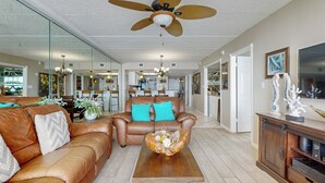 Ein geräumiges Wohnzimmer mit einem braunen Ledersofa, türkisfarbenen Kissen, einem Holztisch und einem Deckenventilator.