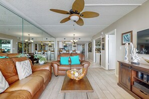 Ein geräumiges Wohnzimmer mit einem braunen Ledersofa, türkisfarbenen Kissen, einem Holztisch und einem Deckenventilator.