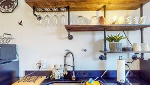 A kitchen with a sink, faucet, and various items like a cutting board, utensils, and a shelf with glasses and mugs.