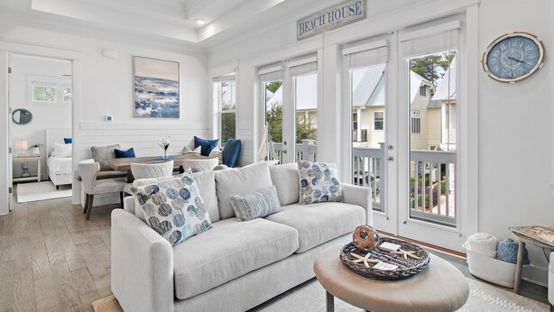 Um interior claro e branco com uma grande janela, um sofá com almofadas estampadas e uma placa com a inscrição 'BEACH HOUSE'.