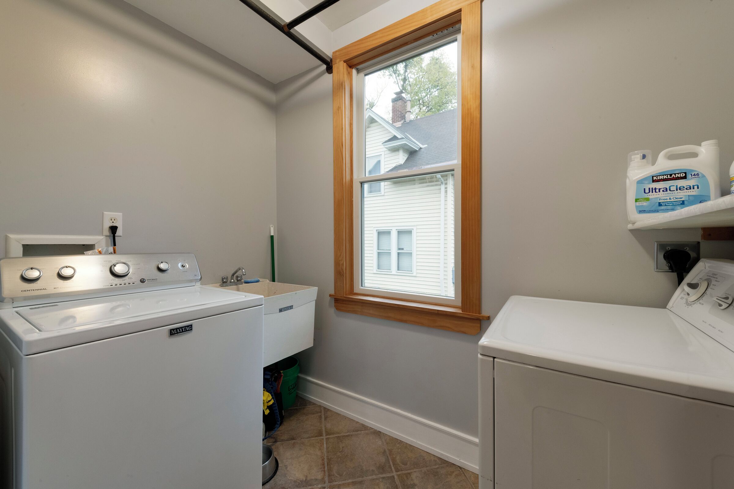 A laundry room with a front-loading washer and dryer, a window with a wooden frame, and a shelf with Kirkland UltraClean detergent.