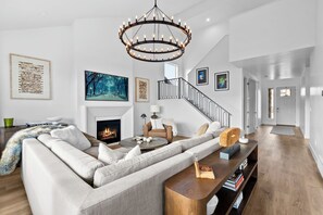 A modern living room with a large sectional sofa, a fireplace, a chandelier, and a staircase. - New Luxury 6BR Mountain Home Minutes from Snowbasin 4,100 Sq Ft w/Mountain Views (Mountain Green)